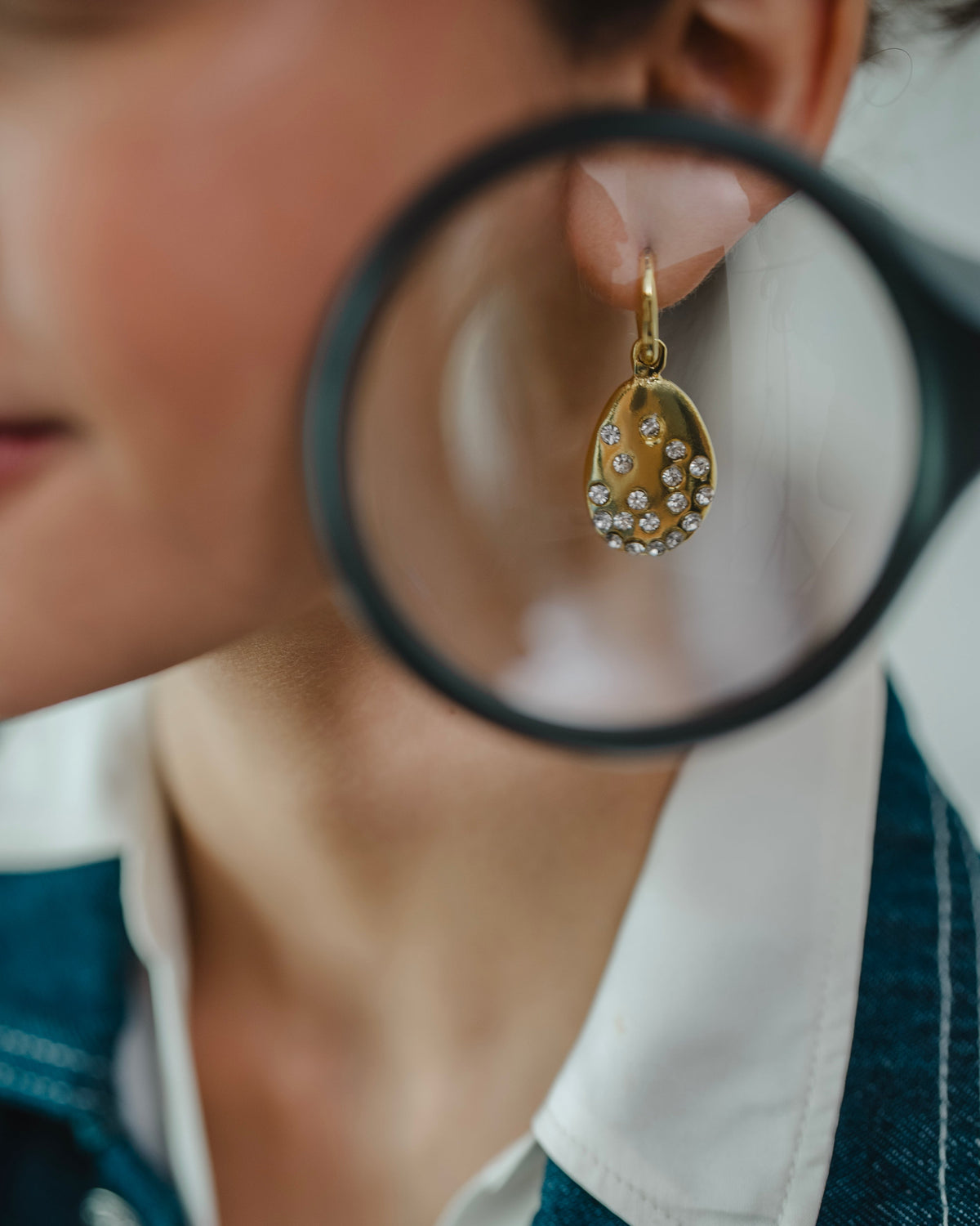 A close-up lifestyle shot of a person wearing a gold teardrop earring adorned with small white pearls, with a black-rimmed magnifying glass held near the earring.