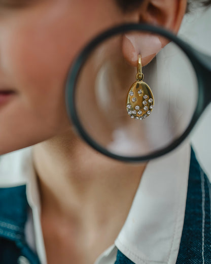 A close-up lifestyle shot of a person wearing a gold teardrop earring adorned with small white pearls, with a black-rimmed magnifying glass held near the earring.